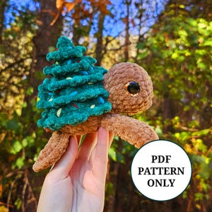 Puede incluir: Un juguete de peluche de tortuga tejido a crochet que lleva un árbol de Navidad verde en la espalda. La tortuga tiene pelo marrón y grandes ojos marrones. El árbol de Navidad tiene luces azules y amarillas.