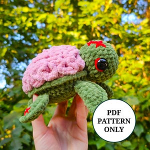 May include: A handmade crochet turtle in shades of green and pink. The turtle has a pink shell, green body, and red accents. The image includes a white circle with the text "PDF PATTERN ONLY".