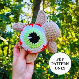 Peut inclure: Une tortue en crochet avec une tête et des nageoires marron clair, dotée d'une carapace en forme de globe oculaire avec une pupille noire, un iris vert clair et une sclérotique blanche avec des détails rouges. Une étiquette indique PDF PATTERN ONLY.