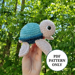 Puede incluir: Un juguete de peluche de tortuga tejido a crochet con una concha azul y verde y un cuerpo beige. La tortuga tiene grandes ojos negros y está siendo sostenida en una mano. El texto "PDF PATTERN ONLY" es visible en la imagen.