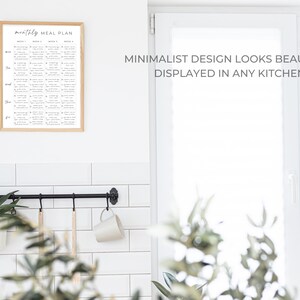 May include: A black metal kitchen utensil rack with hooks hanging on a white tiled wall. A white mug hangs from one hook, and two wooden spoons hang from other hooks. A potted plant sits on a shelf below the rack. A framed printable with the text "monthly meal plan" is hanging on the wall above the rack. The text "MINIMALIST DESIGN LOOKS BEAUTIFUL DISPLAYED IN ANY KITCHEN" is printed on the wall to the right of the rack.