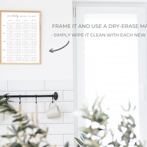 May include: A framed monthly meal planner with a white background and black text. The text says "monthly meal plan" and lists the days of the week and weeks of the month. The image is in a white kitchen with a black metal rack and a plant hanging from it. The text below the image says "FRAME IT AND USE A DRY-ERASE MARKER - SIMPLY WIPE IT CLEAN WITH EACH NEW MONTH".