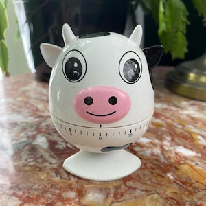 May include: A white cow-shaped kitchen timer with black spots, ears, and eyes, and a pink nose. The timer has a dial with numbers and a base. The timer is on a marble surface.