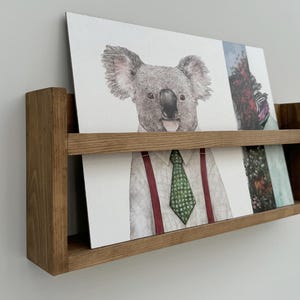 Könnte beinhalten: Ein hölzernes Wandregal mit mehreren Kunstdrucken. Die Drucke zeigen einen Koala mit Krawatte und andere Illustrationen zum Thema Natur. Das Regal ist aus hellbraunem Holz gefertigt und an einer weißen Wand befestigt.