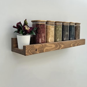 Peut inclure: Une étagère à épices en bois teinté foncé, contenant plusieurs bocaux en verre avec des couvercles en bois. Les bocaux contiennent diverses épices, et un petit pot blanc avec une composition florale violet foncé est à gauche.