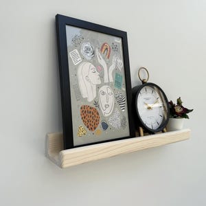 May include: A framed art print with abstract faces and shapes, a black clock, and a small potted plant sit on a light wood shelf. The clock has a black frame and a gold-colored ring at the top. The art print is in a black frame.