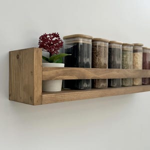 Peut inclure: Une étagère à épices en bois avec une étagère et une petite étagère inférieure. Elle contient plusieurs bocaux en verre avec des couvercles en bois, remplis de diverses épices. Une petite plante en pot avec une fleur rouge est sur le côté gauche.