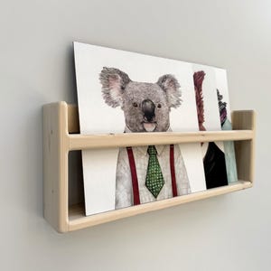 May include: A light-colored wooden wall-mounted shelf holding several art prints. The prints feature animal illustrations, including a koala wearing a tie and suspenders. The shelf is mounted on a light gray wall.