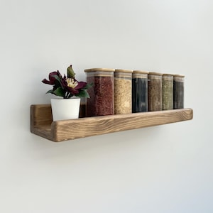 May include: A wooden spice rack with six glass jars filled with various spices. A small white pot with a dark purple flower is on the left. The rack is made of stained wood and is mounted on a white wall.