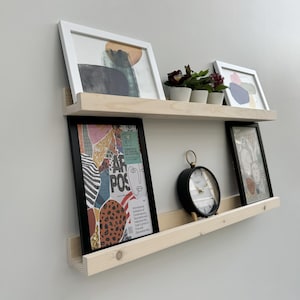 Puede incluir: Estanterías flotantes de madera de dos niveles con obras de arte enmarcadas, pequeñas plantas en macetas y un reloj analógico negro. Las obras de arte incluyen pinturas abstractas y un póster con texto.