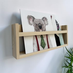 May include: A light-colored wooden wall-mounted shelf holding several art prints. The prints feature animal portraits, including a koala and a person wearing a shirt and tie. The shelf is mounted on a white wall.
