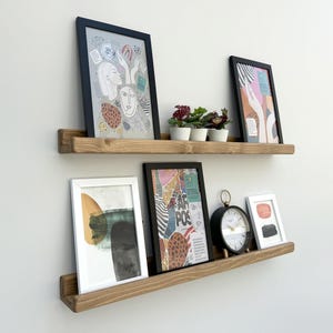 Może przedstawiać: Dwie drewniane półki wiszące z oprawioną grafiką i małymi roślinami doniczkowymi. Górna półka mieści dwa oprawione obrazy i trzy małe rośliny doniczkowe. Dolna półka mieści trzy oprawione obrazy i czarny zegar analogowy.