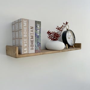 May include: A wooden floating shelf displays decorative items. Three books with titles "La Vie Boheme", "Bohemian", and "Irises" are stacked. A white oval vase holds red branches, and a black clock with a white face sits on the shelf.