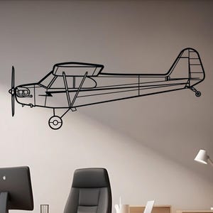 Puede incluir: Arte de pared de avión de metal negro. El avión es un perfil lateral con hélice, alas y cola. El arte está montado en una pared gris claro en un entorno de oficina. La oficina tiene un escritorio, una silla y una computadora.