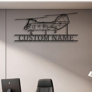 May include: A black metal wall art piece featuring a detailed silhouette of a tandem-rotor helicopter. Below the helicopter, a horizontal bar contains the text CUSTOM NAME in a bold, sans-serif font, integrated into the metal structure.