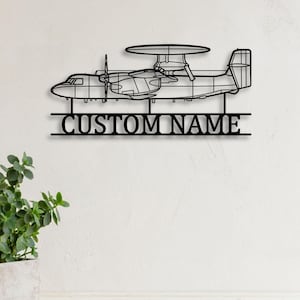 Può includere: Arte da parete in metallo nero raffigurante un aereo con una grande cupola radar. Il testo "CUSTOM NAME" è visualizzato sotto. L'opera d'arte è montata su una parete bianca, con una pianta in vaso in primo piano.