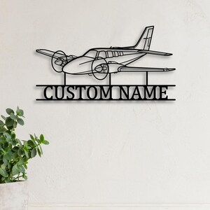 Op de afbeelding: Zwarte metalen vliegtuig wandkunst met de tekst "CUSTOM NAME". Het vliegtuig heeft twee propellers en een gedetailleerd ontwerp. Het kunstwerk is bevestigd aan een witte muur.