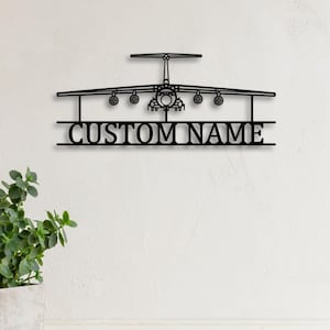 Puede incluir: Arte de pared de avión de metal negro con el texto "CUSTOM NAME" debajo del avión. El avión tiene alas, motores y cola detallados. El arte está montado en una pared blanca, con una planta en maceta en la esquina inferior izquierda.