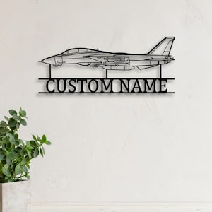 Può includere: Decorazione murale in metallo nero raffigurante un aereo da caccia. Il testo "CUSTOM NAME" è visualizzato sotto l'aereo. L'opera d'arte è montata su una parete bianca, con una piccola pianta in vaso.