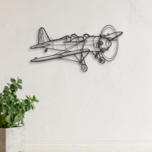 Op de afbeelding: Een zwart metalen vliegtuig wandkunstwerk. Het vliegtuig heeft een propeller, vleugels en een staart, met zichtbare details van de cockpit en het landingsgestel. Het kunstwerk is gemonteerd op een witte muur, met een plant in een pot op de voorgrond.