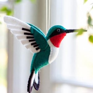 Può includere: Un ornamento a forma di colibrì in feltro, con corpo verde, ali bianche con dettagli neri e gola rossa. L'uccello è sospeso da un sottile filo bianco. Lo sfondo mostra una finestra e del fogliame, creando un'ambientazione naturale.