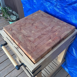May include: A square butcher block with a light wood frame. The butcher block is made of end-grain wood, with a reddish-brown color. The frame has a lower shelf and a handle with black hardware.