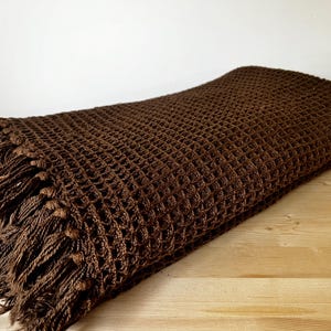 May include: A dark brown woven blanket with a waffle-like texture. The blanket has a fringe along one edge and is folded on a wooden surface. The blanket is a warm, cosy home textile.