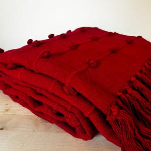Puede incluir: Una manta de tiro roja con un diseño texturizado de pompones y ribete de flecos.