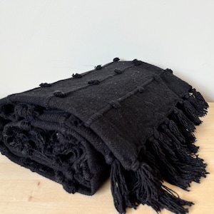 May include: Black throw blanket with a textured woven design and fringe. The blanket is folded and has a soft, cozy look.