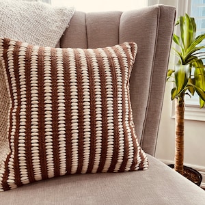 May include: A brown and white striped throw pillow with a textured woven design. The pillow is sitting on a light brown armchair.