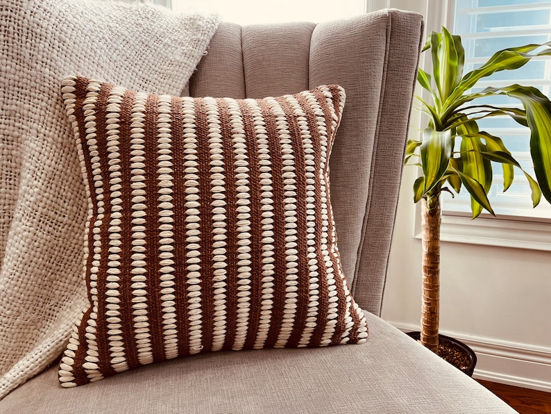 May include: A brown and white striped throw pillow with a textured woven design. The pillow is sitting on a light brown armchair.
