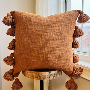 Könnte beinhalten: Ein braunes Kissen mit Quasten an jeder Ecke. Das Kissen liegt auf einem Holzhocker.