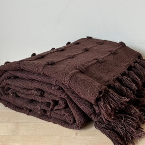 May include: A brown throw blanket with a textured woven design and fringe. The blanket is folded and displayed on a wooden surface.