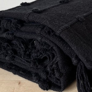 May include: A black throw blanket with a textured woven design and fringe detailing. The blanket is folded and has a soft, cosy appearance.