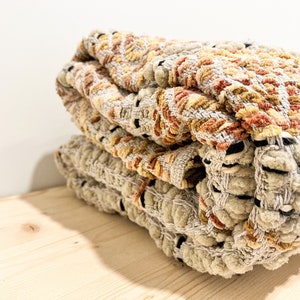 May include: A stack of woven throw blankets in beige, brown, and orange with a textured, fuzzy surface.