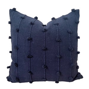 May include: A navy blue decorative throw pillow with a textured design. Vertical lines and small, raised tufts create a pattern across the pillow's surface. The pillow is square-shaped and appears to be made of a soft fabric.