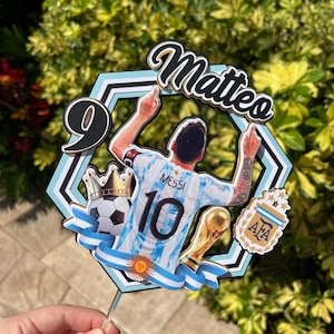 May include: A cake topper featuring a soccer player in an Argentina jersey with the number 10 and the name "Matteo". The design includes a soccer ball, a trophy, and the number 9, all set against a blue and white background.