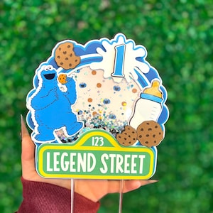 Pode incluir: Um topo de bolo colorido com o Monstro das Bolachas segurando um biscoito, o número 1 e uma mamadeira. O topo tem uma placa de rua que diz "LEGEND STREET" com o número "123". O design inclui biscoitos e glitter.