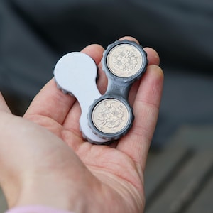 Puede incluir: Un estuche gris y blanco para lentes de contacto que contiene dos monedas. El estuche tiene un diseño único con un asa blanca y dos compartimentos circulares para las lentes. Las monedas son plateadas con diseños intrincados. El estuche se sostiene en una mano.