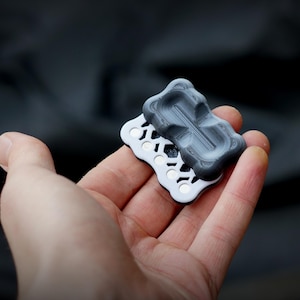 Puede incluir: Dos juguetes fidget entrelazados, impresos en 3D. Uno es gris oscuro y texturizado, el otro blanco con un patrón hexagonal. Se sostienen en una mano sobre un fondo oscuro borroso. El diseño parece ergonómico y mide aproximadamente 5 cm de largo.