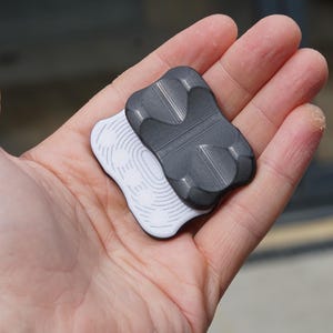 Puede incluir: Dos objetos pequeños de forma abstracta, uno blanco y otro gris oscuro, se sostienen en una mano. El objeto blanco tiene un patrón de líneas curvas, mientras que el gris tiene una superficie texturizada con formas redondeadas. Los objetos parecen estar hechos de un material sólido.