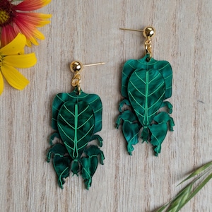May include: A pair of green leaf-shaped earrings with gold-colored posts and ball studs. The earrings have a layered design with detailed veining, resembling real leaves. The background includes yellow and red flowers and a wooden surface.