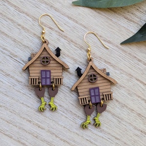 May include: A pair of whimsical wooden earrings. Each earring features a miniature house design with a brown roof, a purple window, and chicken feet dangling below. The earrings have gold-colored ear wires.