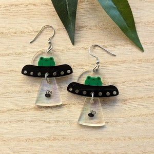 May include: A pair of whimsical earrings featuring a black UFO design with a green frog pilot. The UFOs have a clear, iridescent bottom and a small fly graphic. The earrings are suspended from silver-colored ear wires.