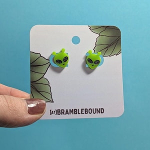 May include: A pair of lime green alien head stud earrings with black glittery eyes, mounted on a white display card featuring green leaf illustrations and the text [AT]BRAMBLEBOUND printed in dark brown at the bottom center of the card.