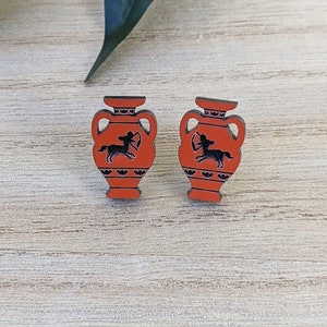 May include: Two red and black earrings in the shape of Greek vases. Each earring has a black centaur silhouette with a bow and arrow. The earrings are displayed on a light wood grain surface.