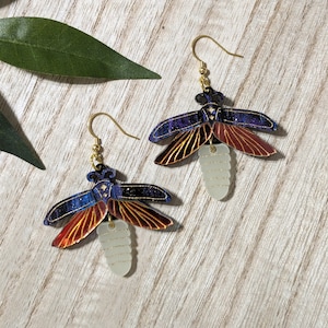 May include: A pair of insect-shaped earrings. The earrings feature a gold hook and a body with blue, red, and gold wings. The lower portion of the insect is a light, translucent color. The earrings are set against a light wood grain background.