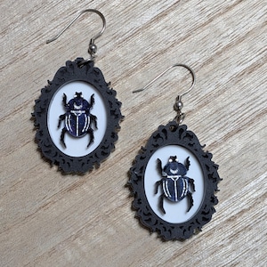 May include: A pair of black framed earrings with a white background, each featuring a dark blue scarab beetle design. The earrings have silver-colored fish hook ear wires and a decorative frame.