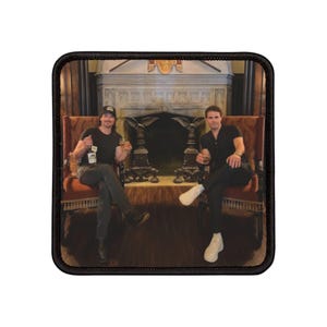 Può includere: Due uomini in camicia nera e jeans sono seduti su sedie ornate di fronte a un camino. Tengono bicchieri in mano e sembrano brindare. Il camino ha un rivestimento in pietra e una grande apertura ad arco. La stanza è arredata in stile tradizionale.