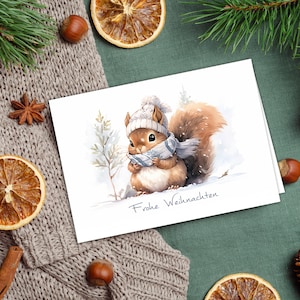 K&ouml;nnte beinhalten: Eine wei&szlig;e Karte mit einer Aquarell-Illustration eines Eichh&ouml;rnchens, das einen wei&szlig;en Strickhut und einen blauen Schal tr&auml;gt. Das Eichh&ouml;rnchen steht im Schnee mit einem Kiefernbaum im Hintergrund. Die Karte sagt "Frohe Weihnachten".